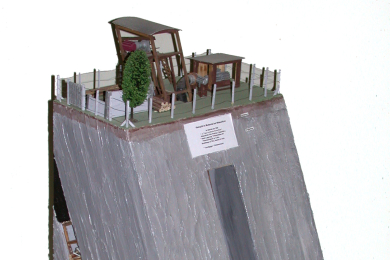 Prismatisch, einseitig abgeschrägtes Modell aus Sperrholz, farbig gefasst, mit Einsichten von drei Seiten in Schacht, Querschlag und Flözstrecke; auf der Oberseite Darstellung der übertägigen Anlagen; Kennzeichnung der Schichten durch Außenbemalung; vorne Schild mit Beschriftung.